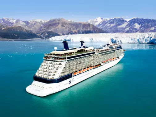 A large Celebrity cruise ship with multiple decks sailing through Hubbard Glacier, surrounded by icy blue waters and rugged mountainous terrain