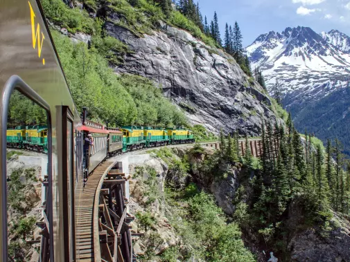 A scenic train in Skagway crossing a wooden trestle bridge in a lush mountain landscape. with rocky cliffs on one side and tall pine trees under a clear blue sky.