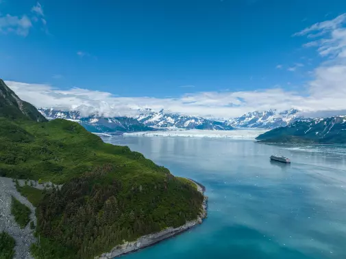 A serene view of Hubbard Glacier with a lone cruise ship, and distant snow-capped mountains under a bright blue sky.